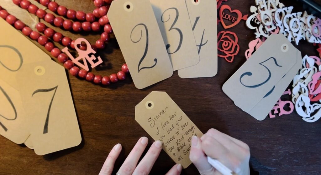 Handwriting a Valentines Day love note on numbered brown craft tags on desk for family